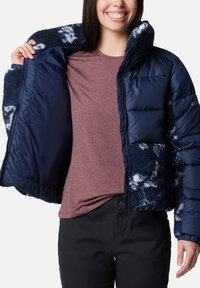 Giacca a vento blu navy con fodera interna in pile e maniche a fantasia; presenta colletto alto e chiusura con zip; indossata sopra una camicia bordeaux.
