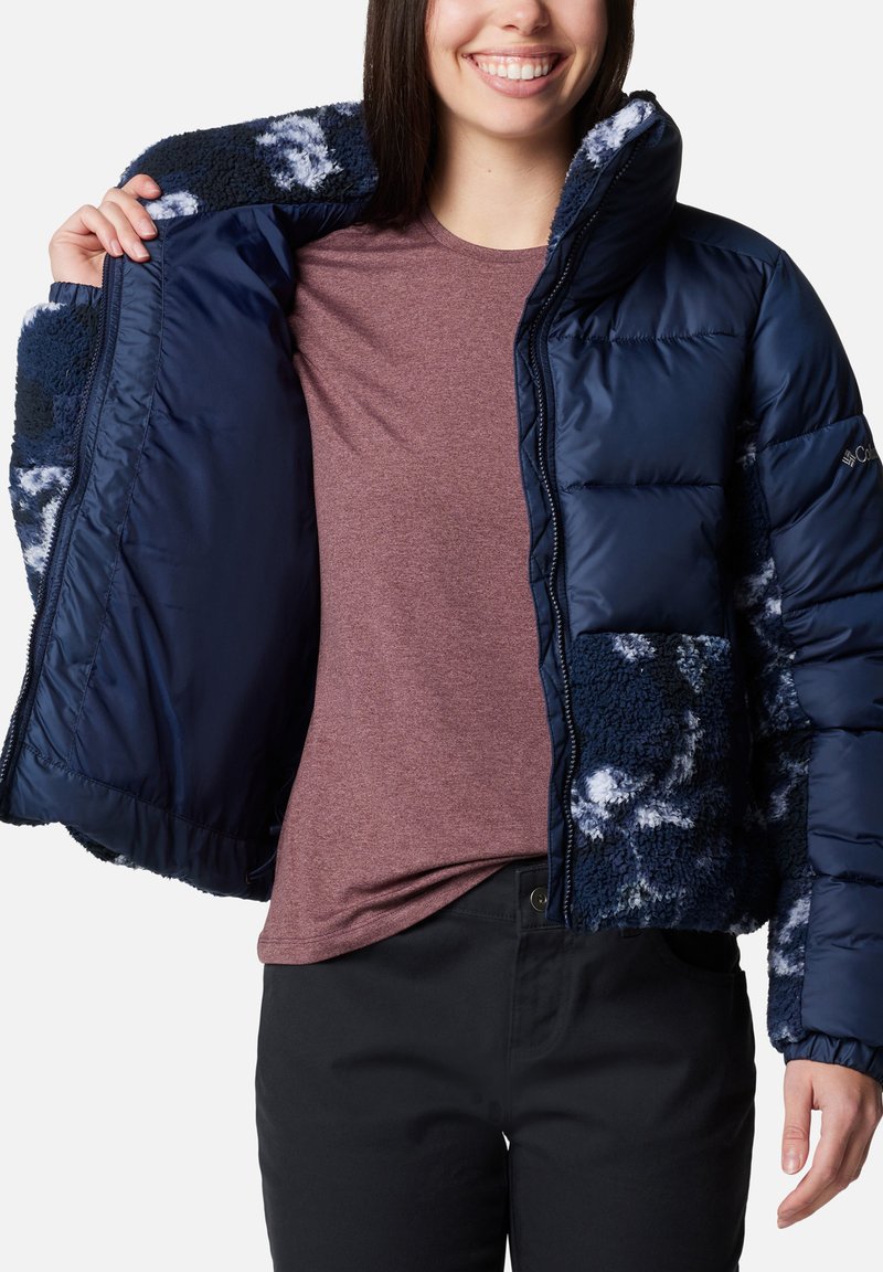 Doudoune marine avec doublure en polaire et manches à motifs ; équipée d'un col haut et d'une fermeture éclair ; portée par-dessus une chemise bordeaux.