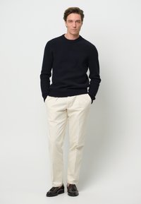 Marinblå stickad tröja med ribbad textur, krämfärgade byxor och mörkbruna skor. Modellen står med händerna i fickorna.