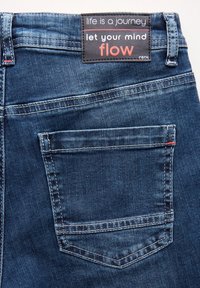 Mörkblå denimjeans med en bakficka med kontrasterande vit sömnad. Etikett ovanför fickan har röd och vit text på en svart bakgrund.