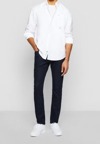 Biała koszula z długim rękawem, w połączeniu z ciemnoniebieskimi dżinsami i białymi sneakersami. Koszula ma małe logo na piersi.