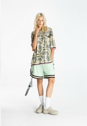 Persona rubia de pie, con camisa de gran tamaño con estampado de cachemira, pantalones cortos verde menta con ribete negro, calcetines blancos y zapatos beige sin cordones.