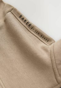 Zbliżenie na beżowy materiał z przeszytą metką z czarnym tekstem "BAKER BY TED BAKER" na off-white tle.