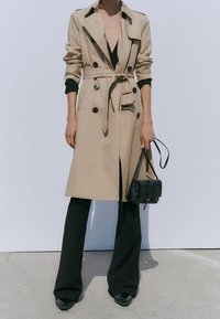 Trench-coat beige à boutons croisés, taille ceinturée et col à revers, associé à un pantalon évasé noir et un petit sac à main noir.