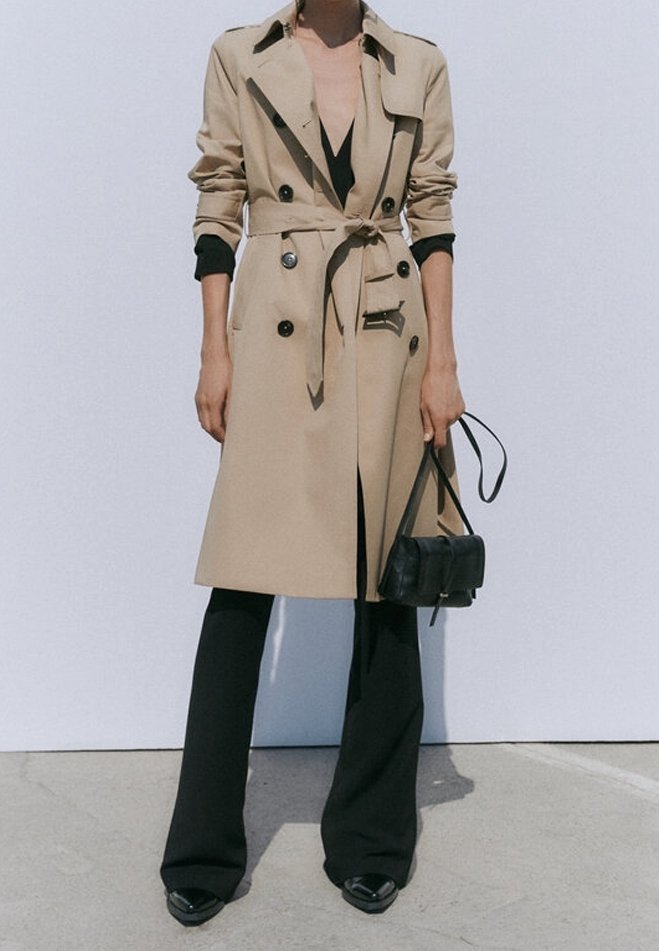 Trench-coat beige à boutons croisés, taille ceinturée et col à revers, associé à un pantalon évasé noir et un petit sac à main noir.