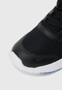 Chaussure de sport noire avec un dessus en mesh, semelle en caoutchouc, embout en cuir lisse et système de laçage. Des accents incluent un élément de design bleu.