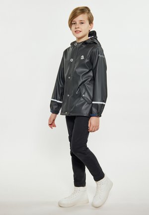 Zwarte waterdichte jas met een capuchon, voorzien van witte reflecterende strepen en drukknoopsluiting, gecombineerd met zwarte broek en witte hoge sneakers.