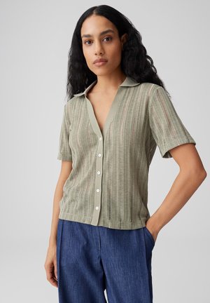 Frau mit langen lockigen Haaren, die ein hellgrünes kurzärmeliges Hemd mit Knöpfen und blaue Hosen trägt, steht mit einer Hand in der Tasche.