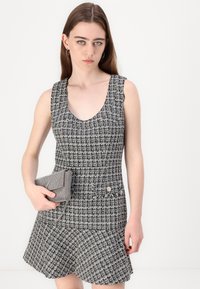 Musta-valkoinen ruudullinen mekko, jossa tiukka yläosa ja levenevä hame. Rakenteellinen kangas, hihaton malli ja koristeelliset napit. Pitelee samannäköistä käsilaukkua.