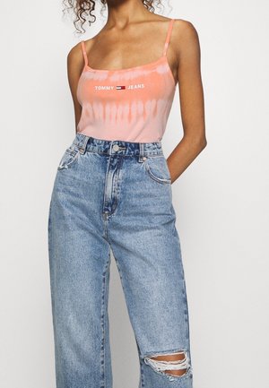 Femme portant un débardeur tie-dye rose avec le logo "Tommy Jeans" et un jean bleu taille haute avec un genou déchiré sur fond blanc.