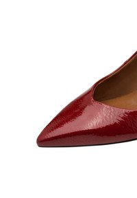 Bout pointu d'une chaussure à talons aiguilles rouge brillante avec une finition en cuir texturé et un doublure intérieure beige clair sur fond blanc.