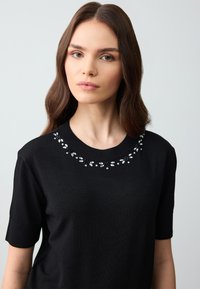 Zwarte trui met korte mouwen, voorzien van een versierde halslijn met doorzichtige kralen en decoratieve stiksels, gemaakt van zachte, gestructureerde stof.