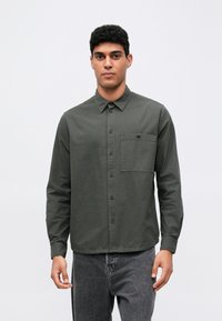 Camisa de manga larga, de color verde oscuro, con cuello abotonado, bolsillo en el pecho y una tela texturizada. Combinada con jeans grises. Diseño sencillo.