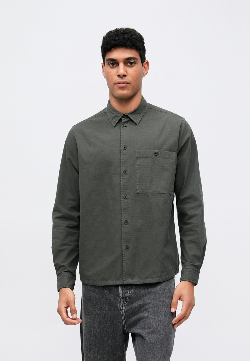 Camisa de manga larga, de color verde oscuro, con cuello abotonado, bolsillo en el pecho y una tela texturizada. Combinada con jeans grises. Diseño sencillo.