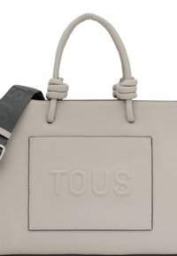 Sac fourre-tout texturé beige avec des poignées nouées et une sangle grise amovible. Présente le logo "TOUS" en relief sur une poche avant.