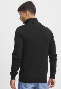 Schwarzer gestrickter Pullover mit hohem Kragen, gerippten Bündchen und Saum, ausgestattet mit einem strukturierten Muster und lockerer Passform, kombiniert mit hellblauen Jeans.