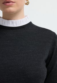 Zwarte trui met een ronde halslijn, voorzien van een gerimpelde witte kraag. Zachte textuur en aansluitend ontwerp met minimale zichtbare naden.