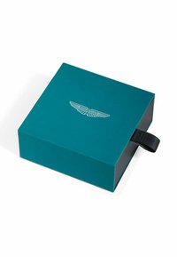 Boîte en carton bleu sarcelle avec un intérieur noir. Présente un logo Aston Martin blanc sur le dessus et une languette en tissu noir pour l'ouverture.