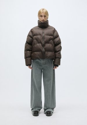 Bruine pufferjack met hoge kraag, ritssluiting en grote baffles. Gecombineerd met grijze denim broeken met wijde pijpen en zwarte schoenen.