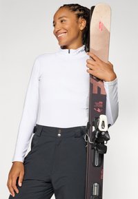Smilende kvinne holder et ski med bindinger, iført en hvit zip-up genser og svarte skibukser, satt mot en enkel lys bakgrunn.