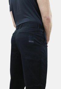 Pantaloni neri realizzati in un misto cotone; le caratteristiche includono una tasca posteriore con etichetta, passanti per cintura e un design a gamba dritta.