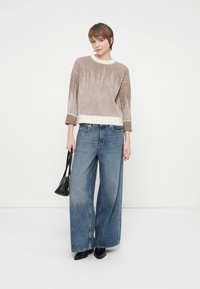 Kvinde iført en beige og hvid strikket sweater, bredbenede blå jeans, sorte spidse sko og holder en sort håndtaske foran en hvid baggrund.