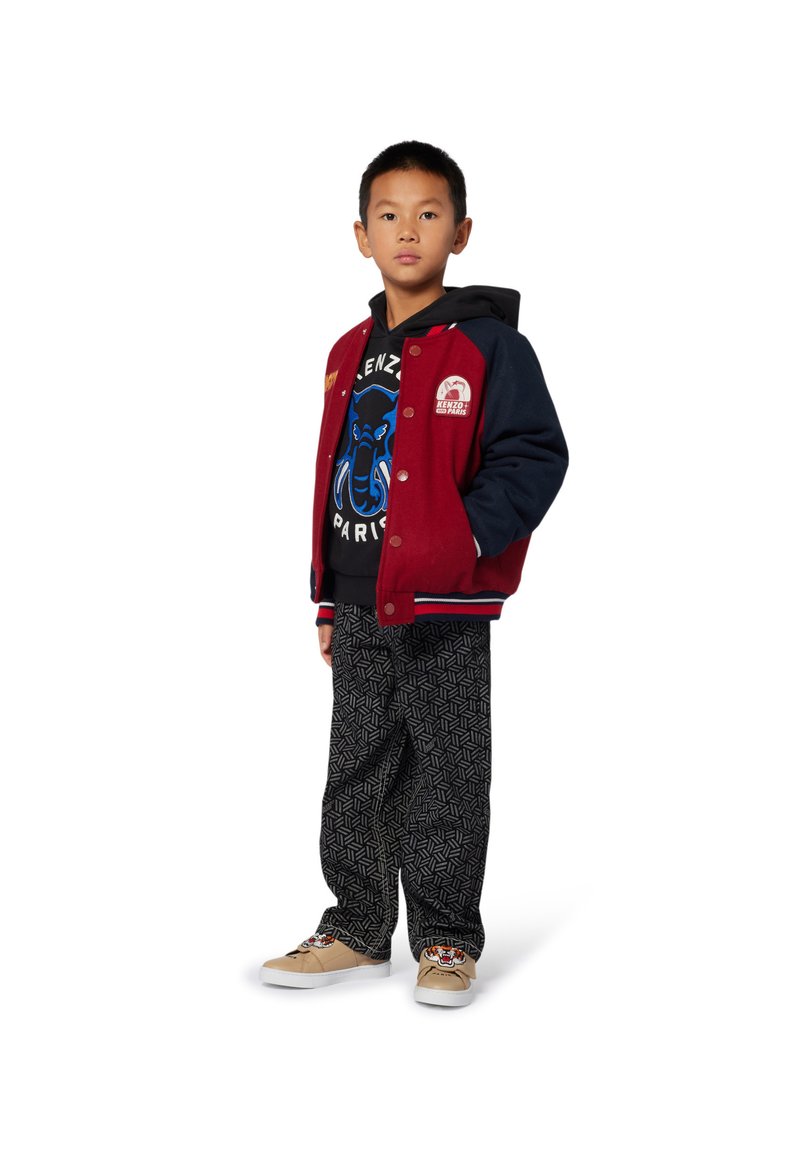 Giacca varsity rossa e navy con dettagli bianchi; felpa nera con grafiche blu; pantaloni neri stampati; scarpe beige con ricami.