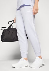 Ανοιχτό λευκό παντελόνι φόρμας με λευκές λεπτομέρειες και λογότυπο, σε συνδυασμό με λευκά αθλητικά παπούτσια, κρατώντας μια μαύρη αθλητική τσάντα με λαβές.