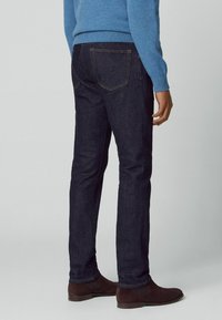 Dunkelblaue Jeans aus Denim mit Slim-Fit, ausgestattet mit klassischen Gesäßtaschen und sichtbaren Nähten. Getragen mit braunen Stiefeletten.