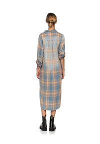 Blå rutig lång klänning med uppvikta ärmar, med orangea och gula accenter. Materialet verkar mjukt; modellen bär svarta ankelskor.