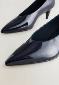 Escarpins en vernis bleu marine avec un bout pointu et un design épuré. Présente un talon modeste et une texture lisse avec une finition brillante.