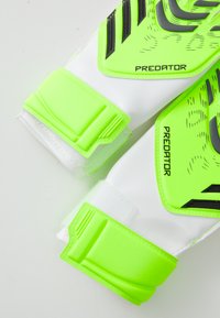 Mănuși de portar verzi și albe cu accente negre. Prezintă o zonă de prindere texturată și curele Velcro pentru o fixare sigură. Inclusiv marca "PREDATOR".
