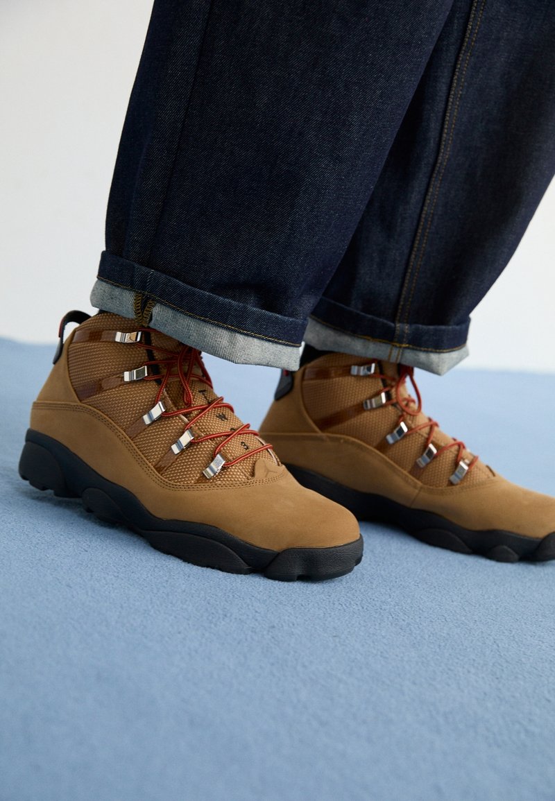 Braune Wanderschuhe mit einem Obermaterial aus Mesh und Wildleder, roten Schnürsenkeln, silbernen Hardware-Highlights und einer schwarzen Gummisohle. Kombiniert mit dunklen Denim-Jeans.