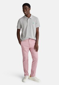 Šedá polo triko s zeleným logem, spárované se světle růžovými kalhotami a bílými teniskami. Oblečení má neformální design vhodný pro teplé počasí.