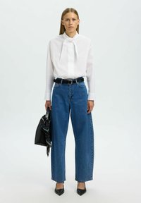 Blusa blanca con cuello de lazo, vaqueros de talle alto azules, cinturón negro y tacones negros de punta afilada. Sosteniendo un bolso negro con un pañuelo a rayas.