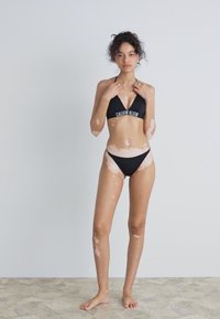 Črn bikini komplet z trikotnim zgornjim delom in nizko rezanimi hlačkami. Model ima srednje temno polt in valovite lase, stoji na strukturirani svetli površini.
