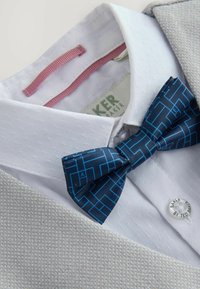 Giacca di abito grigia chiara con una camicia bianca sotto. Un papillon blu navy con un motivo geometrico e un dettaglio in nastro rosa.