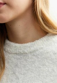 Ljusgrå, texturerad tröja med hög, ribbad halsringning. Mjuk, fluffig material med små fläckar. Långt, rakt brunt hår synligt.