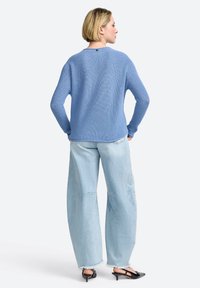 Blauer Rippenstrickpullover mit langen Ärmeln, rundem Ausschnitt und subtilen Nahtdetails auf der Rückseite. Kombiniert mit hellen, weit geschnittenen Jeans.