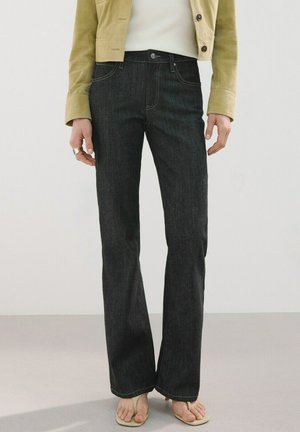 Kobieta ubrana w ciemne dżinsy o prostych nogawkach, jasnobeżową krótką kurtkę, biały top i beżowe sandały z odsłoniętymi palcami, stojąca na jednolitym tle.