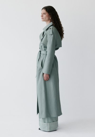 Kvinde med krøllet hår iført en lang, bæltebindet trenchcoat i salviegrøn og matchende bukser med vide ben, stående i profil mod en ensfarvet baggrund.