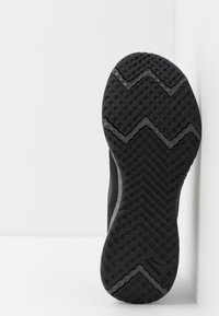 Semelle de chaussure de course noire avec un motif de semelle en zigzag, positionnée à la verticale contre un mur blanc dans un coin.