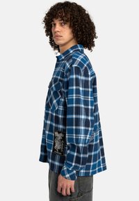 Blå og marineblå skovmandsskjorte i flannel med lommer, der har et sort patch med hvid grafisk detalje på siden og knappede manchetter.