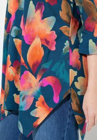 Tunic floreale in verde teal scuro con grandi fiori arancioni, rosa e turchesi. Orlo irregolare con spacchi laterali. Tessuto morbido e leggero.