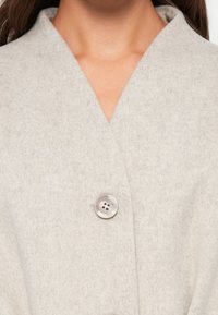 Blazer in lana grigia chiara con colletto a V, chiusura con un singolo bottone metallico e tessuto morbido e testurizzato.