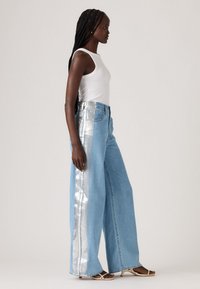 Woman with long braids wearing white sleeveless top, wide-leg blue jeans with silver side stripes, and beige open-toe heels, standing sideways.