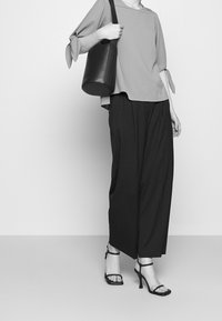 Blusa grigia chiaro con maniche annodate, pantaloni neri a gamba larga e tacchi neri con cinturini; porta una borsa rotonda in pelle nera.