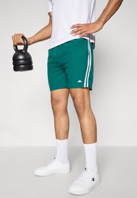 Žalios sportinės šortai su baltais šonų juostelėmis, derinami su baltais sportbačiais ir iki kelių siekiančiomis kojinėmis. Vienoje rankoje laikomas juodas kettlebell.