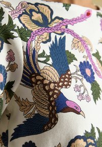 Wzorzysta tkanina z wielokolorowym ptakiem z niebieskimi i różowymi detalami, otoczona kwiatowymi motywami w zieleni, różu i granacie na białym tle.