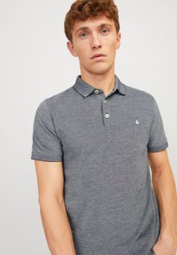 Collared, short-sleeve polo shirt in dark gray fabric with a textured pattern, featuring three buttons and a small logo on the chest.